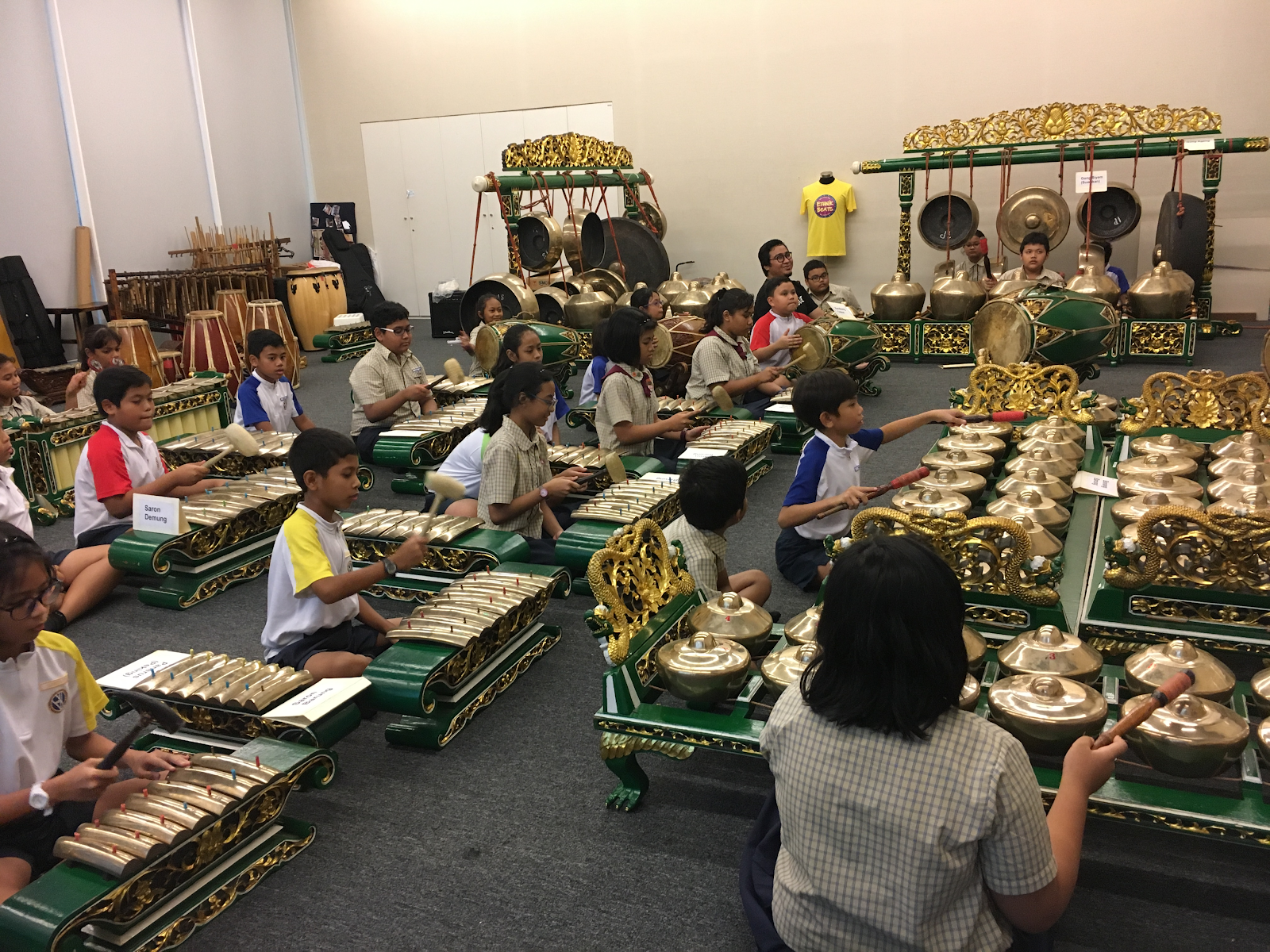 Children sit on the floor playing traditional gamelan instruments with gold and green decorations in an indoor room.
